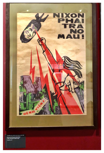 Photo of a framed vintage poster on a museum’s red wall, with an illustration depicting a woman standing in a bombed-out village. She carries a dead child in one arm and defiantly raises her fist to a falling bomb bearing Richard Nixon’s face. Red lightning bolts stretch between the woman and the bomb — perhaps indicating the bomb’s destructive power or (my preferred idea) the woman’s rage, the rage of a people, striking back. Large all-caps text on the poster reads “Nixon phải trã nợ máu!” (“Nixon must pay a blood debt,” translated on the museum placard as “Nixon owes the Vietnamese a debt of blood”).
