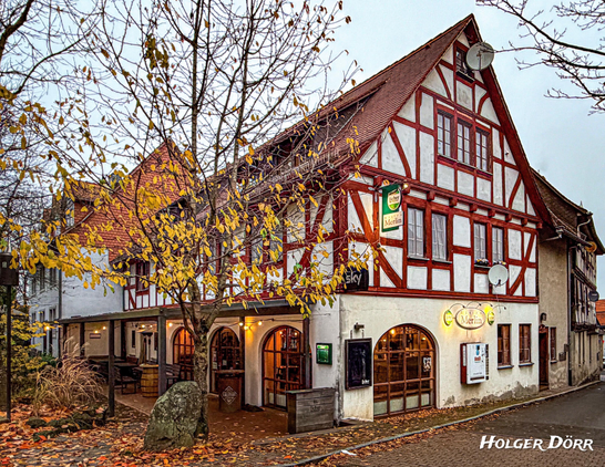Fachwerkhaus mit rotem Gebälk und weißem Putz in der Altstadt von Grünberg, Hessen. Im Erdgeschoss leuchtet das Bistro Merlin, während herbstliche Blätter auf das nasse Kopfsteinpflaster fallen. Stimmungsvoll beleuchtete Szene an einem kühlen Abend.