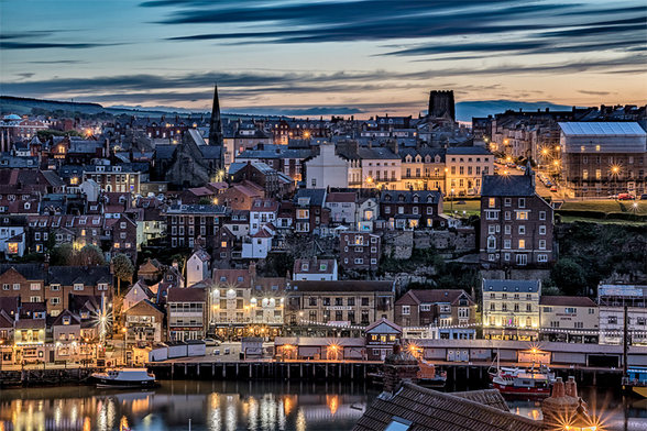 alt-text please

This is a stunning twilight photograph of Whitby, a historic coastal town in North Yorkshire, England. The image captures the town's iconic seaside character with its distinctive layers of architecture climbing up the hillside from the harbor.

In the foreground, fishing boats are moored along the waterfront, their lights reflecting beautifully in the still harbor waters. The town's buildings rise dramatically up the cliff, showing a rich mix of traditional Georgian and Victorian architecture in varying shades of brick and stone. Street lights twinkle throughout the scene, creating a warm, inviting glow against the deepening dusk sky.

The buildings are tightly clustered in the characteristic style of historic English coastal towns, with narrow streets winding between them.

The sky features dramatic streaked clouds in shades of deep blue and peach, creating a moody, atmospheric backdrop. The composition beautifully captures that magical "blue hour" moment when artificial lights begin to dominate but natural light still illuminates the scene, giving the photograph a romantic, timeless quality that showcases Whitby's enduring charm as one of England's most picturesque harbor towns.


