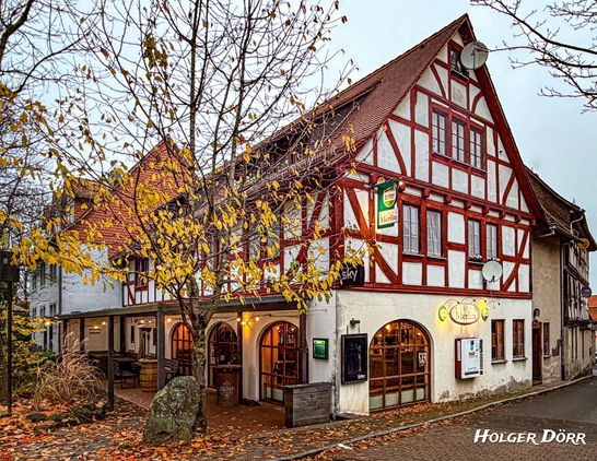 Fachwerkhaus mit rotem Gebälk und weißem Putz in der Altstadt von Grünberg, Hessen. Im Erdgeschoss leuchtet das Bistro Merlin, während herbstliche Blätter auf das nasse Kopfsteinpflaster fallen. Stimmungsvoll beleuchtete Szene an einem kühlen Abend.