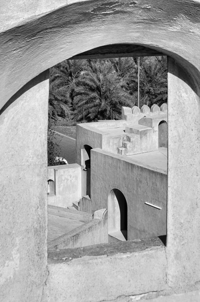 Eine Schwarz-Weiss-Aufnahme, die durch einen gewölbten Bogen auf eine terrassenartige, orientalisch geprägte Architektur blicken lässt. Die klar strukturierten Mauern und Bögen verleihen der Szene eine geometrische Harmonie. Im Hintergrund sind Palmen zu erkennen, die den orientalischen Charakter des Ortes unterstreichen. 