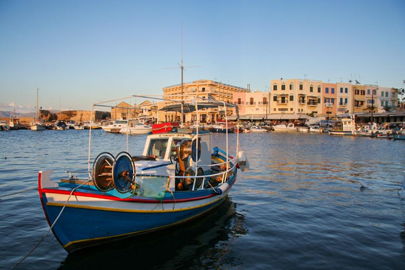 A colorful fishing boat is moored in a harbor, surrounded by various boats and buildings with Mediterranean architecture. The scene is illuminated by warm sunset lighting, reflecting off the water.