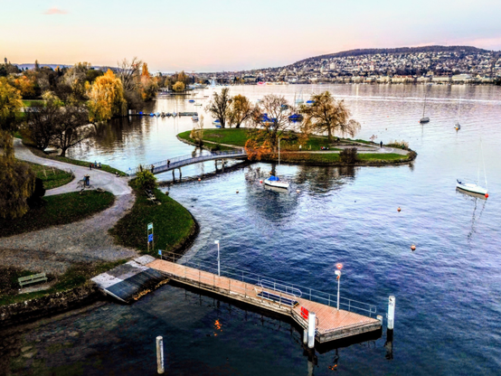 An aerial shot of Lake Zurich with Zurich in the far distance, autumn-colored trees in the midground and the lake with a jetty in the foreground 