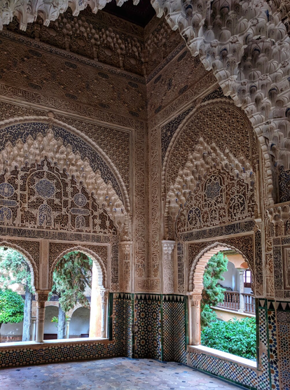 An interior photograph of an intricately designed historical architectural space within the Alhambra in Granada, Spain. The image showcases a richly decorated room featuring Islamic architectural elements, such as horseshoe arches and elaborate stucco work. The walls and ceilings are adorned with detailed geometric patterns, arabesques, and calligraphic inscriptions, all meticulously carved into the plaster. The arches frame views of a lush courtyard filled with greenery, including trees and plants, which contrast beautifully with the earthy tones of the architecture. The floor is composed of patterned tiles, adding to the overall aesthetic harmony of the space. The lighting highlights the depth and intricacy of the carvings, creating a serene and majestic atmosphere.