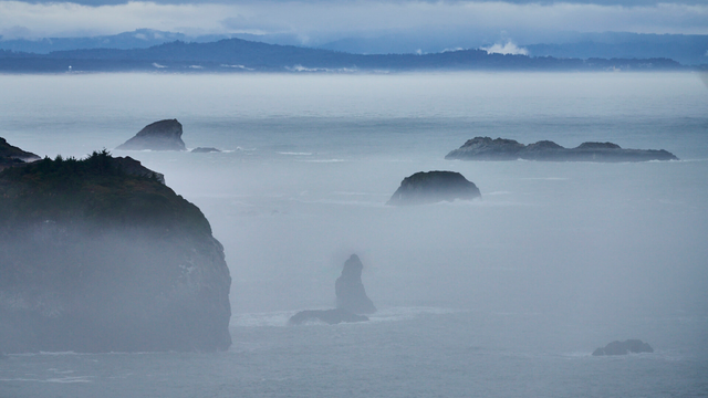 Offshore rocks and sea stacks are rise through a low level fog. Clouds and fog shroud distant hills.