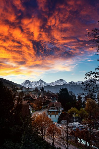 Ein atemberaubender Sonnenaufgang über einem malerischen Dorf, umgeben von majestätischen Bergen. Der Himmel ist spektakulär gefärbt mit intensiven Rottönen, die sich in den Wolken spiegeln. Die Häuser des Dorfes liegen friedlich in der Landschaft, während im Vordergrund Bäume und Sträucher in herbstlichen Farben zu sehen sind.