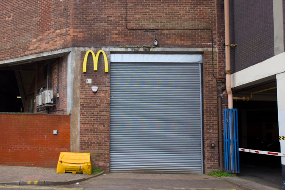 A large metal roller shutter is set into a brick building. Above the door is a yellow McDonald’s M logo, but there are no other restaurant signs. Nearby are a yellow bin, a red brick wall, and a blue gate leading to a car park.