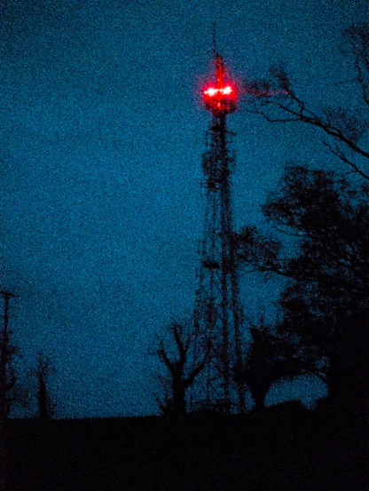 A photograph of a radio tower at night, red lights illuminating the top part of the tower. few trees are seen, Dark Blue sky. all foreground elemnts are black.