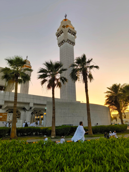 A low-angle photograph capturing a large white mosque with a distinctive geometric minaret reaching into an orange and yellow sunset sky. Lush green bushes and palm trees line the foreground, where several individuals dressed in white robes are relaxing or moving about.