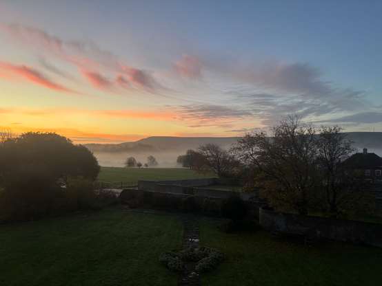 Sunrise from Glynde. A few whispy clouds lit from the the left by the glow of the soon-to-rise Sun. Firle Beacon in the distance. The garden in the foreground. Mist enveloping trees near Glynde Reach in the middle ground. 