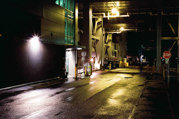 A service road runs between tall buildings. There are bridges, pipes, and ducts between the buildings so that it looks almost subterranean.