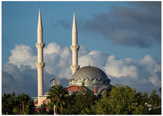 A skyline featuring a mosque with two tall minarets and a large dome, surrounded by green trees and a backdrop of blue sky with fluffy clouds.