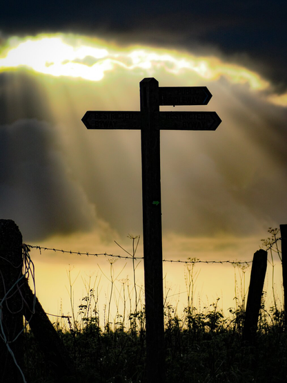 Silhouette of a fingerpost, behind it fingers of sunlight through a dark cloud.