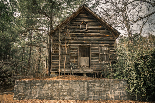A weathered two-story wooden cabin with horizontal wood siding sits elevated on a stone foundation wall in a wooded setting. The structure shows significant age and deterioration, with darkened, graying wood planks throughout. The cabin features a steep pitched roof, a small attic vent window near the peak, a central doorway with a damaged door, and windows on either side of the entrance. A wooden porch or deck spans the front. Young bare trees grow close to the building, and the cabin is surrounded by a mix of evergreen and deciduous trees. A neat stone retaining wall made of rectangular gray blocks runs along the front at ground level. The forest floor is covered with fallen leaves and pine needles, giving the scene an abandoned, rural atmosphere.