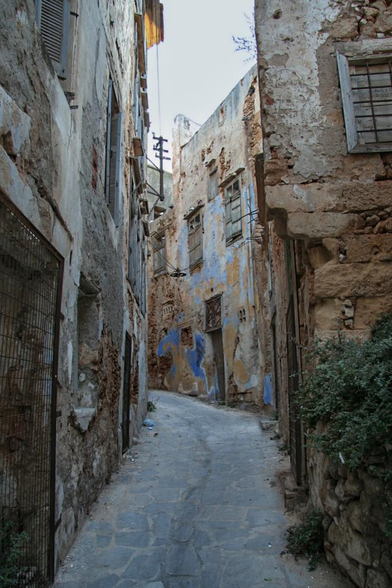 A narrow, winding alleyway flanked by weathered buildings featuring peeling paint and wooden shutters. The ground is paved with stones, and the scene conveys a rustic, historic ambiance, with a few plants visible amidst the urban decay.