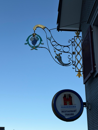 Ein dekoratives Restaurantschild verfügt über ein Design mit Traubenmotiven, mit einem stilisierten Vogel- und Weinrebenelementen, die vor einem klaren blauen Himmel stehen. Darunter steht ein kreisförmiges Schild mit der Aufschrift "Feldschlösschen RESTAURANT TRAUBE".