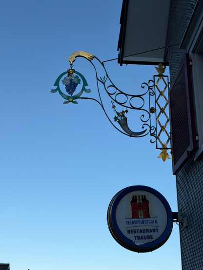 Ein dekoratives Restaurantschild verfügt über ein Design mit Traubenmotiven, mit einem stilisierten Vogel- und Weinrebenelementen, die vor einem klaren blauen Himmel stehen. Darunter steht ein kreisförmiges Schild mit der Aufschrift "Feldschlösschen RESTAURANT TRAUBE".