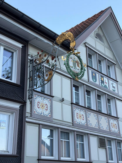 An einer Gebäudefassade hängt ein dekoratives Schild mit einem goldfarbenen Vogel auf einem kreisförmigen Emblem. Das Gebäude hat eine Mischung aus traditionellen und modernen architektonischen Elementen, mit farbenfrohen Zierdesigns unter den Fenstern. Der Himmel ist klar und blau.