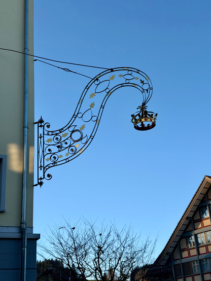 Ein kunstvolles Metallschild mit fließendem Design mit Blättern und einer dekorativen Krone hängt an einem Gebäude vor einem klaren blauen Himmel. Im Hintergrund gibt es kahle Bäume und einen Teil eines Gebäudes mit einem traditionellen architektonischen Stil.