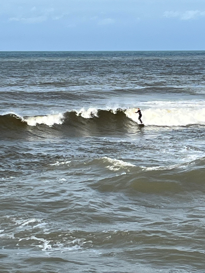 A lone surfer rides the edge of a breaking wave. A tumultuous grey-green sea after the storm, under a blue sky 