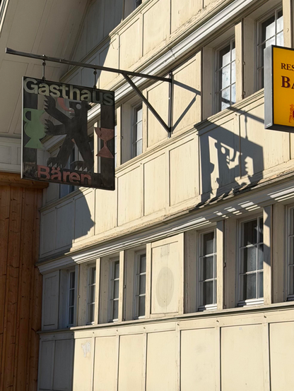 Ein Schild für "Gasthaus Bären" hängt an einem Gebäude mit einem Bären und Symbolen, die Tassen ähneln. Der Hintergrund zeigt eine cremefarbene Holzfassade mit mehreren Fenstern. Das Schild wirft einen Schatten auf die Wand und verstärkt die architektonischen Details.