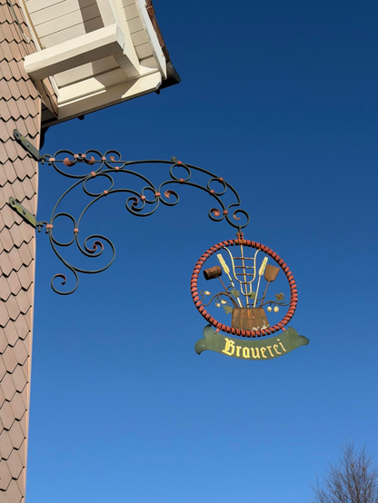 Ein dekoratives Schild, das an einem Gebäude hängt, mit einem kreisförmigen Design mit Brauwerkzeugen und dem Wort "Brauerei" vor einem klaren blauen Himmel.