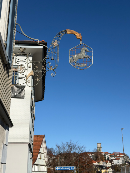 Ein dekoratives Metallschild mit einer Pferdesilhouette hängt an einem Gebäude, mit komplizierten Schriftrollen und einer vogelähnlichen Figur darüber. Der Hintergrund zeigt einen klaren blauen Himmel und Dächer von nahe gelegenen Gebäuden. Ein Straßenschild mit der Aufschrift "Mittelbissauweg