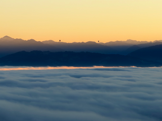 Eine ruhige Landschaft mit rollenden Wolken unter einem bergigen Horizont bei Sonnenuntergang. Silhouetten-Heißluftballons sind zu sehen, wie sie über den Gipfeln vor einem abgestuften Himmel in warmen Orange- und Blautönen schwebt.
