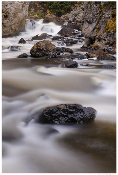 Long-exposure photograph of Firehole Falls in Yellowstone National Park, showing smooth, flowing water cascading over dark rocks between rugged canyon walls, surrounded by moss and scattered autumn foliage