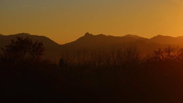 Evening photo of a hilly landscape tinted orange-yellow by the light from the setting sun. In the background is a forest-covered ridge with a castle ruin on one of the hills. In the front is a valley with some trees.
