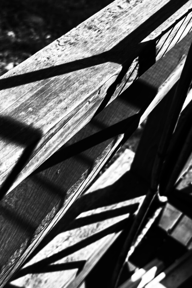 The photograph shows a close, angled view of weathered wooden boards crossed by sharp, high-contrast shadows. The sunlight carves deep black geometric shapes across the planks, turning the railing or structure into an abstract pattern of intersecting lines. The extreme contrast and skewed perspective make the scene feel tense and slightly disorienting, as if the wood and shadows are folding into each other without a stable horizon