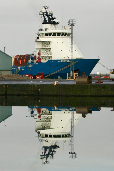 A blue boat moored in a dock in the top half of the photo with a perfect reflection of it on the water in the bottom half 