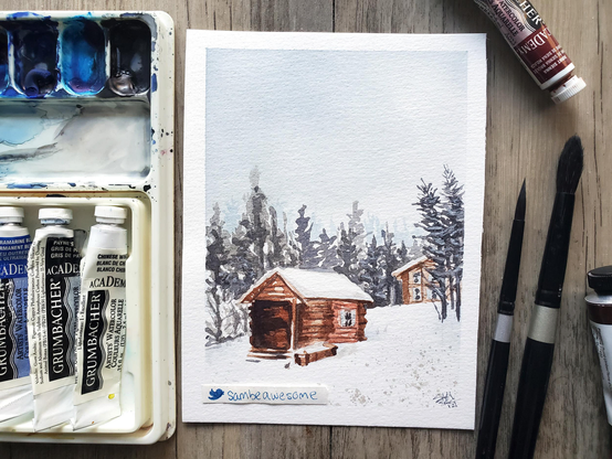 A photo of a watercolor painting of a wooden shed, blanketed in snow, with a forest behind it, and a cabin peeking out from the distant trees. There are splatters of silver paint for an iridescent look that looks very pretty in person. Next to the painting are brushes and paints.