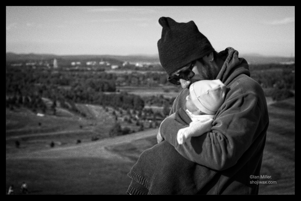 A man in a beanie and sunglasses holds a baby wrapped in a blanket close to his chest, surrounded by a scenic view of hills and trees in the background. The image is in black and white.
