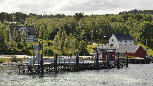 A photo of a small ferry terminal in front of a hill. The hill is covered in forest and has a couple houses on it. The sky is filled with clouds.
