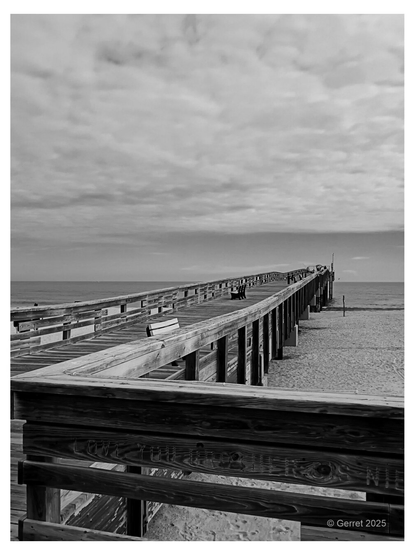 Black and white image of a long wooden pier extending out to sea under a cloudy sky. The scene is serene and slightly melancholic.