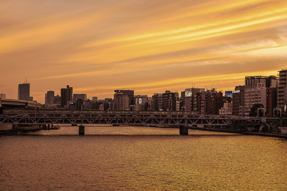 Tokyo Asakusa cityscape at sunset, with high buildings and a train crossing a bridge over a golden Sumida river, under a beautiful orange sky ornated with horizontal strips of clouds beautifully brushed by a light wind. A few neon commercials typical to Japanese big cities are visible on the tops on some building.