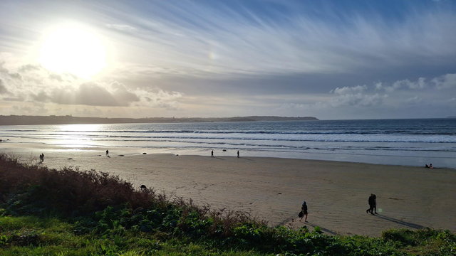 Un paysage de bord de mer en contre-jour. Le soleil en haut à gauche se détache au dessus des nuages. Sur le reste du ciel bleu les nuages s'étirent.
La mer est calme avec quelques vagues au bord. 
Sur la plage on aperçoit des personnes qui se promènent.
Au 1er plan, un peu d'herbe
