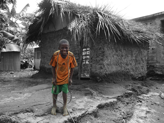 Un enfant en t-shirt et short orange est debout, souriant, devant une habitation d’un village de montagne à Anjouan