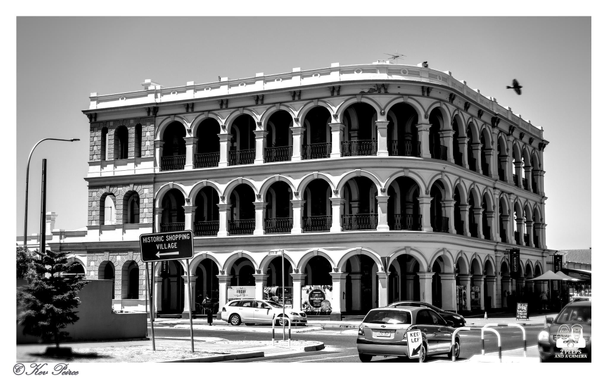 A black and white photograph of the historic three story Largs Bay Hotel in South Australia, featuring a striking facade with arched verandas on the second and third floors.

The architecture is detailed with numerous repeating arches and small balconies with balustrades.

In the foreground, there is a street corner with a few parked cars, and a traffic sign points to a "HISTORIC SHOPPING VILLAGE" to the left. A bird is visible in flight over the rooftop on the right side.