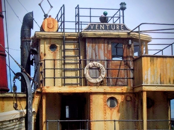 The close-up view of the bridge and cabin of a weathered, rusty steamship movie prop named "VENTURE" painted in large black letters above the wheelhouse.

The cabin is clad in stage aged wood and oxidised yellow-brown metal, featuring several portholes, a ladder leading to a top deck railing, a life preserver hanging on the front railing, and a large black smokestack partially visible on the left.

The overall appearance suggests a neglected, hard-working tramp steamer from the 1930s.

It was docked at the old Miramar Wharf awaiting sailing out to sea for long shots which, apparently, was a dodgy bit sailing as the ocean tried to sink it.