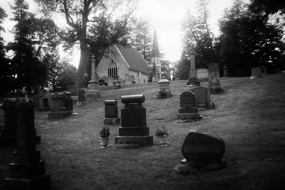 Looking up a small hill in a cemetery towards a small building with a tower. Headstones for various shapes and sizes can be seen as well as large trees in this black and white image.