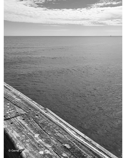 Black and white photo of a calm ocean view from a weathered wooden pier, with scattered clouds in a wide sky, evoking a serene, contemplative mood.