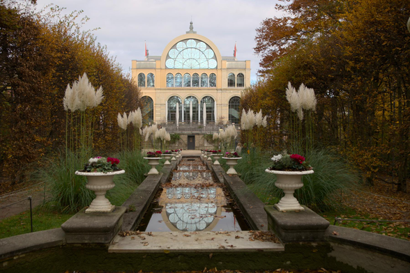Auto-generated description: A formal garden features a series of planters with flowers and tall grasses leading to a grand building with large arched windows and flags.