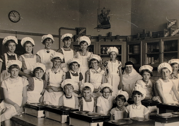 A black and white photograph of a group of approximately 20 young girls posing in a kitchen classroom. All of the girls are wearing white caps and white aprons over their clothing. The teacher (a nun) is visible in the middle row toward the right.

The background features cabinets or shelves, some with glass doors, containing various items which appear to be canned or packaged goods. A round wall clock is visible on the upper left wall, showing the time is 10:45.