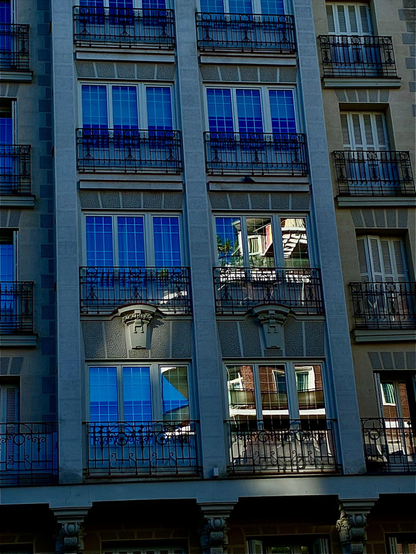 Fachada gris con ventanales en los que se refleja el edificio que hay enfrente, iluminado por el sol.