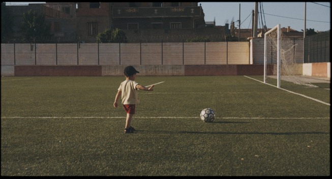 Child on a soccer field pointing at a ball, wearing a yellow shirt and red shorts, with buildings in the background.