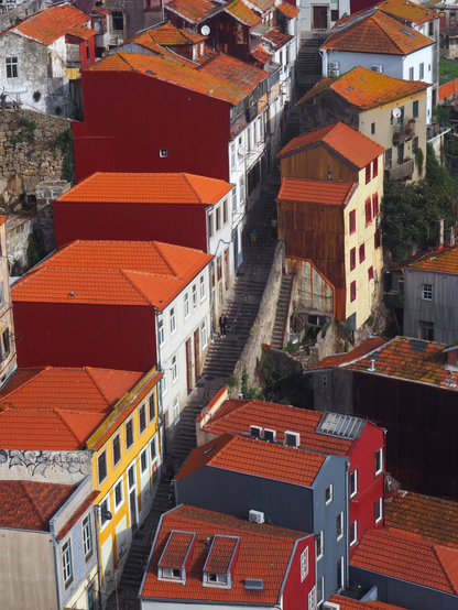 Picture of buildings with red roofs that are built in a hill right next to each other in two rows. Between the buildings there goes really long stairs up the hill. Because of a shadow these stairs look a dark line  in the photo going from bottom left towards upper right. There is also some people walking up and down the steps. Picture has been taken at a higher level.