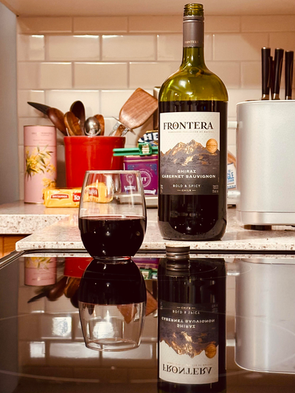 A bottle of Frontera Shiraz Cabernet Sauvignon next to a more-than-half-empty glass of the red wine, on a reflective black stovetop.