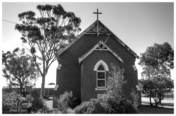 A black and white photograph of a small, rustic brick church, identified by a sign on the gable as "CATHOLIC CHURCH." The building has a simple arched window and a cross atop the peaked roof.

It is framed by large, mature gum trees on the left and various shrubs in the foreground, creating a contrast between the architecture and the Australian bush setting.